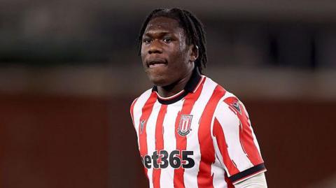 Sydney Agina standing on the pitch with his arms by his side, wearing a Stoke City red and white striped home shirt