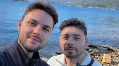 Joe Diaz and Dan Campbell. Both have brown hair. Joe is wearing a black jumper and Dan a white jumper. They are standing in front of a large body of water. 