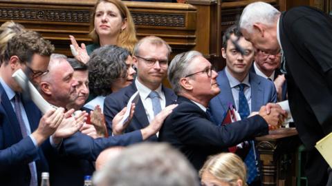 Keir Starmer shakes hands with Lindsay Hoyle, Speaker of the House of Commons, as people clap and pat Stamer on the back after his last PMQ session as Prime Minister