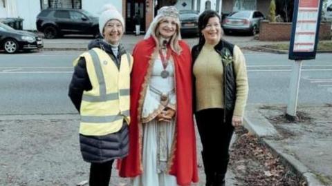 Three women in a row. The one in the centre is dressed in the white dress and red robe of Lady Godiva. 