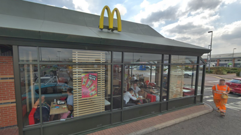 The front of McDonald's in Greenbridge Retail Park, Swindon. The golden arches M is on top of the building and there are people sitting inside at tables. There is a man in high vis walking along the side, holding a McDonald's bag and the car park is visible in the background. 