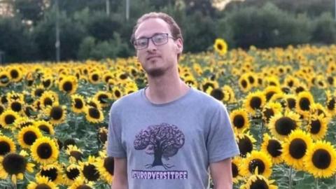 A man with short brown hair and black rectangular rimmed glasses is standing in a field of sunflowers and wearing a grey t shirt with a tree on it with the word neurodiversitree underneath it.