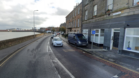 Cars on Trinity Crescent in Edinburgh. There are flats on the right and the sea on the left behind a wall.  