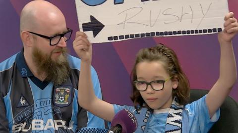 A bald headed man with a long beard wearing an old Coventry City shirt with a Coventry City scarf around his neck sat next to a boy wearing a Coventry City t shirt and scarf around his neck whilst holding up a poster with a black arrow pointing to the word Rushy which is above a dotted line. They are in a BBC studio with a microphone that says BBC CWR and are sitting against a purple background.
