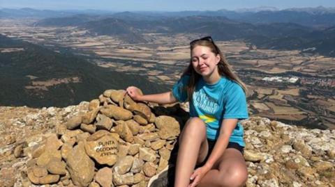A young woman with long blonde hair, a blue tshirt and dark shorts sitting on a pile of brown rocks with a landscape in the distance behind her