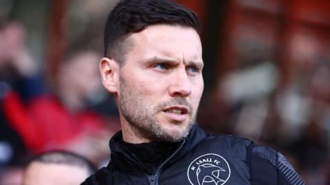 Walsall manager Mat Sadler watching a game, wearing a dark tracksuit