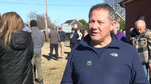 Chris Hems gives a TV interview in a gravelled area. He has greying hair and wears a dark blue half-zipped top. Fellow protesters are standing alone and in groups.