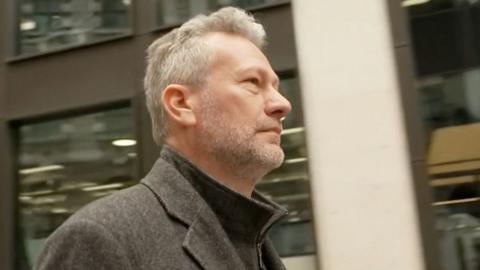 A side shot of Nathan Gill. He has a grey jacket on and a grey stubbly beard. He has short grey hair. 