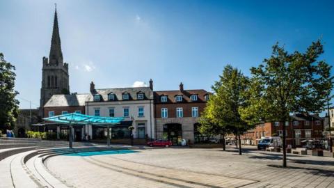 Kettering Market Square, showing several trees, a church, a shelter, buildings surrounding the square and cars in the distance. 