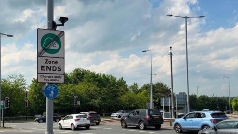 A busy urban road junction on a partly cloudy day. In the foreground, there is a tall grey pole positioned slightly to the left of the center. Attached to this pole is a rectangular road sign with a green circular symbol that contains a cloud-like shape and the letter “C” A diagonal black line crosses through the symbol, indicating the end of a designated zone. Beneath the symbol, the sign clearly reads “Zone ENDS” in bold black letters, followed by smaller text stating “Charges apply Pay online”. Below this sign, there is a smaller circular blue sign showing a white bicycle and a pedestrian, indicating a shared path for cyclists and pedestrians.