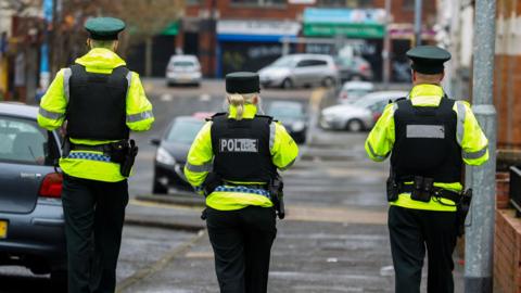 Three police officers are walking down a street in Belfast. There are two males and a female in the middle. They are wearing full police uniform including hats, bullet proof vests and high-visibility jackets. 