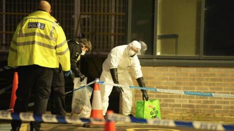 Police tape cordons off the scene, with two forensics officers picking things up from the floor.