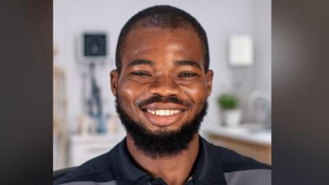An AI-generated image of a man smiling. He is in a kitchen which is blurred in the background. He has a medium-length, dark beard and short dark hair. He's wearing a dark blue and grey polo shirt.