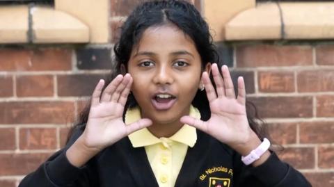 A young girl wearing school uniform has her hands by her face as if she is yelling