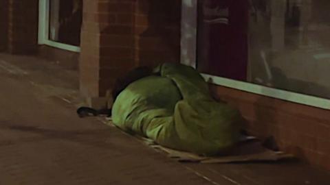A rough sleeper in a green sleeping bag sleeps beside a row of shop windows
