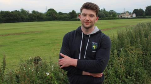 A man - Councillor George Simmonds - standing with his arms folded in front of him wearing a black hoodie and looking straight-faced with a green field in the background. 