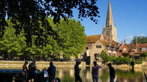 A group of people chat beside a boat, by a river. Sunshine illuminates a church in the background.