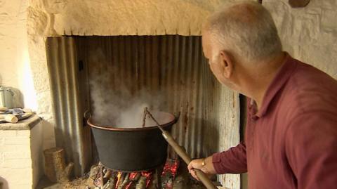 A man wearing a burgundy polo shirt holds a long stick out towards a large copper pan above a log fire and stirs the contents inside it.