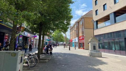 A picture of Staines high street, with trees, benches, bike railings, people and shops.