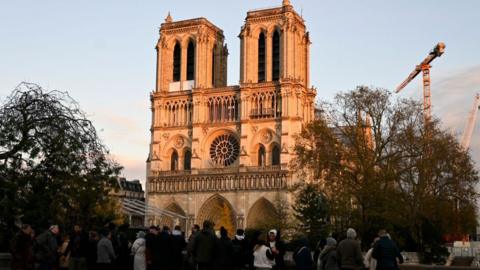 Wide shot of Notre Dame cathedral as it approaches the end of restoration work. A large number of people stand in the foreground and a crane stands behind the building. There are trees either side of the cathedral.  