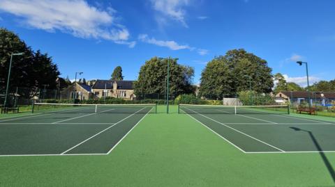 Two tennis courts pictured on a sunny day with trees and houses in the background