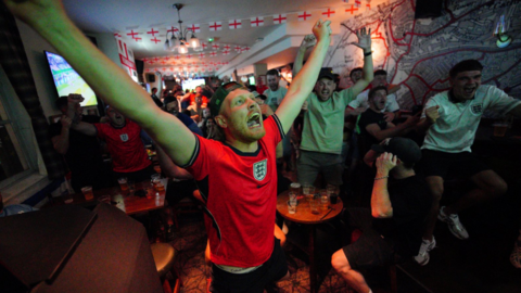 A close-up of a fan wearing a red England shirt, with his arms outstretched in the air and his mouth open with joy. Other fans are celebrating similarly in the background of the pub room, which has England flag banners strung from the ceiling