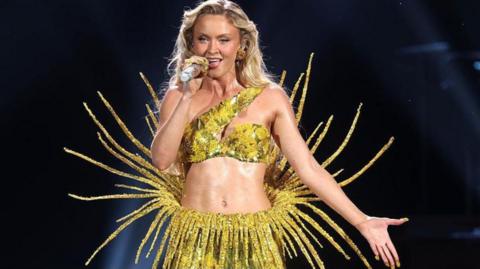 Zara Larsson wearing a yellow sparkly two piece outfit. She has long blonde hair and she is singing into a microphone.