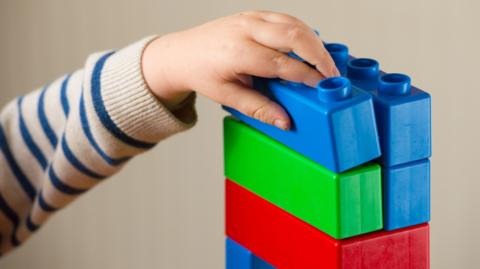 The hand of a young child wearing a blue and white stripped jumper placing a large blue children's building block on top of a stack of other blue, green and red blocks.