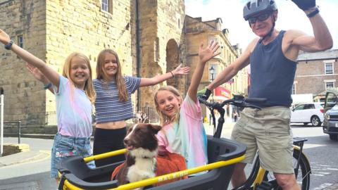 A picture of a man, with a child and a dog in a wagon attached to the front of his bike, and two children standing alongside.