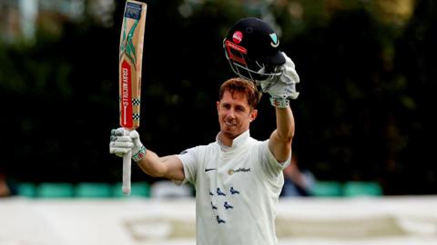 John Simpson holds his bat and helmet in the air to acknowledge his century