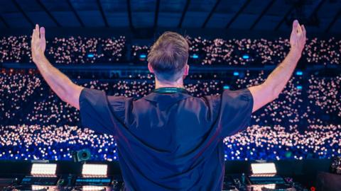 Calvin Harris stands with his back to the camera raising his arms to the crowd of people in front of him.