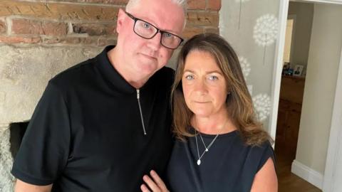 Gary and Angela Daulby look at the camera with their arms around each other in their home. Gary has very short white hair and is wearing black framed glasses. Angela had shoulder length brown hair. Both are wearing black tops