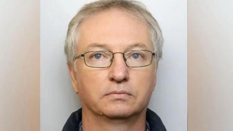 A custody picture of Chapman. He has no expression on his face, and wears dark, thin-framed glasses. 