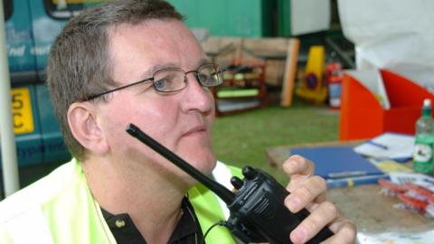 The image shows a person wearing a high-visibility yellow safety vest over a dark shirt. The man is holding a black two-way radio close to his mouth. The background appears to be an outdoor setting with green structures and various items scattered on a table, including a blue folder, papers, and a green bottle. There are also some orange and yellow objects in the distance, possibly safety equipment or barriers.