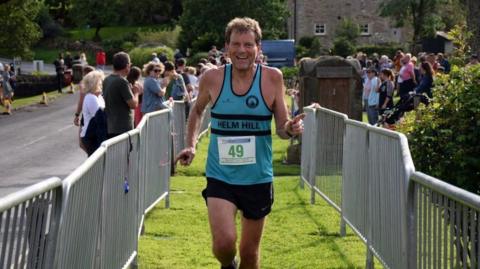 David Griffin, a 66-year-old man, is running towards the camera. He is wearing a blue vest which has the number 49 on it and is running between silver metal barriers which suggest the end of a race. In the distance a number of people are watching the runners and applauding.