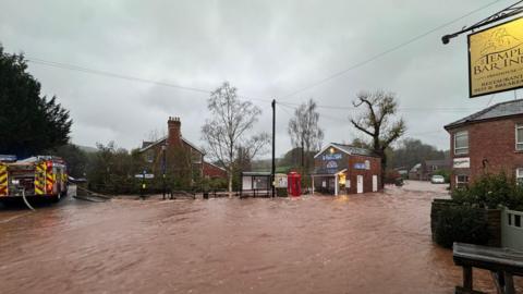 Dark orange floodwater running through a village. The water has covered the road and is sweeping through the street. There is a pub on the right and a bus stop, phone box and fish and chip shop that are all flooded. A fire engine is on the left trying to pump out the water.
