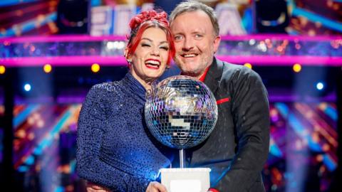 Dianne Buswell and Chris McCausland standing together, smiling and holding the glitterball trophy after winning the Strictly Come Dancing final