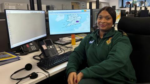 Monet Lobman-Edghill sits in the London Ambulance Service control room. She is wearing a green quarter-zip jumper with 'NHS' and a gold embroidered crest. She is sitting at a desk with two computer screens, a keyboard, a phone and a headset. There is a map of the UK on one of the computer screens. 