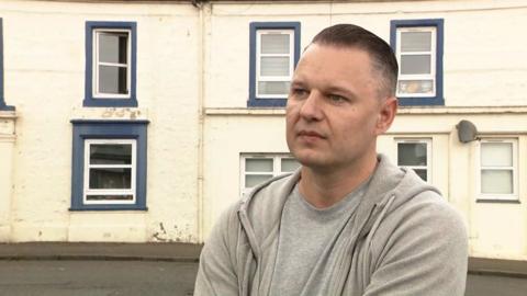 William Podwalski has short hair, shaved at the side, he wears a grey T-shirt and grey hoodie