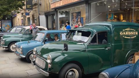Classic cars and small vans in blue and green lined up along a street outside shops. They are faced outwards, on display. 