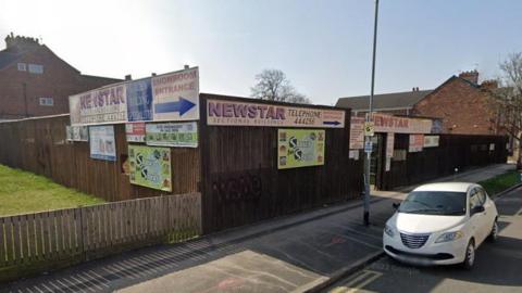 A Google Street View image of the shed supplier, with signs advertising the business on a fence outside. There is also a lawn and cars parked outside