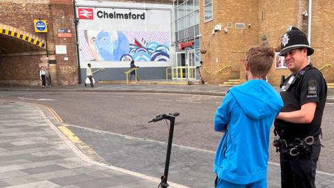 A police officer talking to a person wearing a blue tracksuit and trainers. They are standing by an E-scooter. They are stood by Chelmsford railway station in Essex