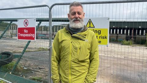 A man is standing in front of a metal fence. He has a beard and is wearing a green coat. No trespassing signs are on the fence.