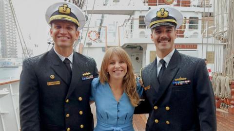 Two naval officers wearing smart uniforms and white military hats stand in the rain either side of a BBC reporter wearing a blue jumpsuit, on the wooden deck of a tallship, with ropes wound neatly behind them 