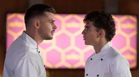A man with short dark hair and a shorter man with short dark hair, both wearing white shirts, facing each other in front of pink and yellow patterned light