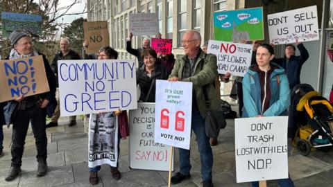 A group of men and women stand outside Cornwall Council's HQ to protest a planning proposal. Several have placards voicing their disgust at the plans. One of the signs near the front of the crowd has "community not greed" written in blue block capitals.