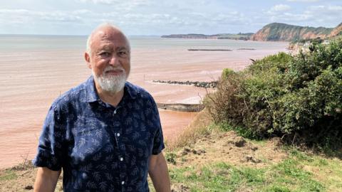 Paul Griew, with a grey beard and a blue short-sleeved shirt, standing at the end of his clifftop garden, close to the edge of a cliff, with the sea beyond 