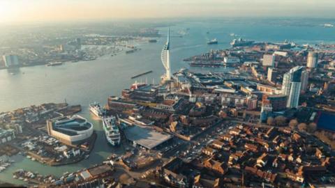 Aerial view of Portsmouth with waterfront around housing and buildings with the Spinnaker Tower on the edge of the city and numerous ships on the water.