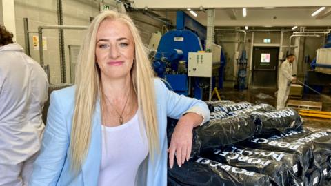 A woman with long blonde hair, a light blue blazer and a white top stands in a factory, with an elbow resting on a pile of bags of porcelain clay. Behind her, one man in a white overall is spraying the factory floor with water, another is working at a machine. A large blue machine and fire exit can also be seen in the background.