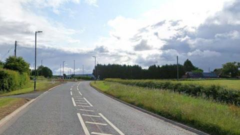 A main road, with grass verge and hedge on the left and a field on the right. A roundabout can be seen in the distance and a national speed limit sign on a lamppost on the left. There are buildings in the far distance.